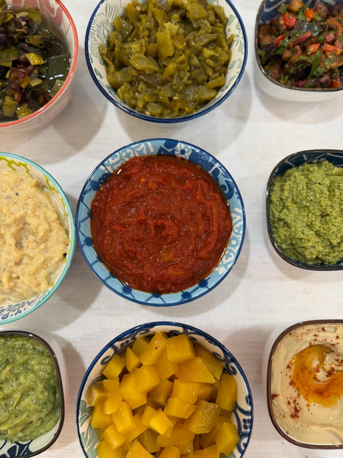 Overhead view of nine bowls of Israeli dips including matbucha, pesto, hummus and yellow beets