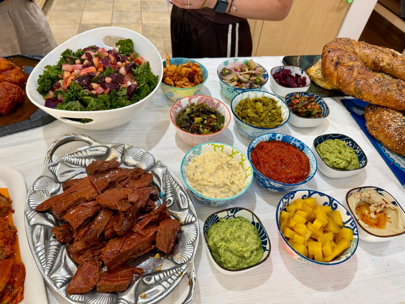 A full table with corned beef, kale salad, dips and seeded challahs